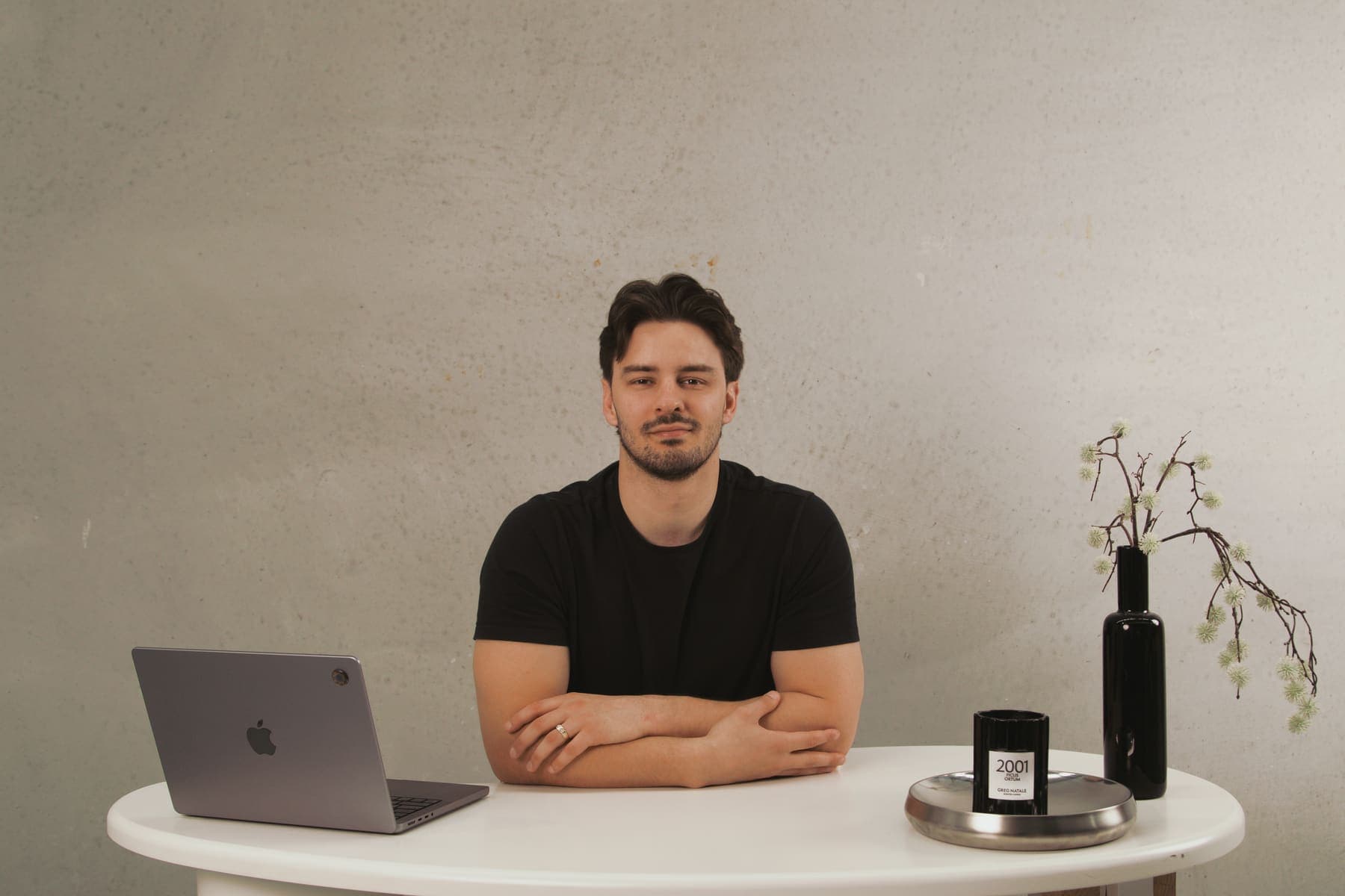 Luke Marinovic at a white desk with his arms folded on the table, laptop open, a black vase of blossom beside him.