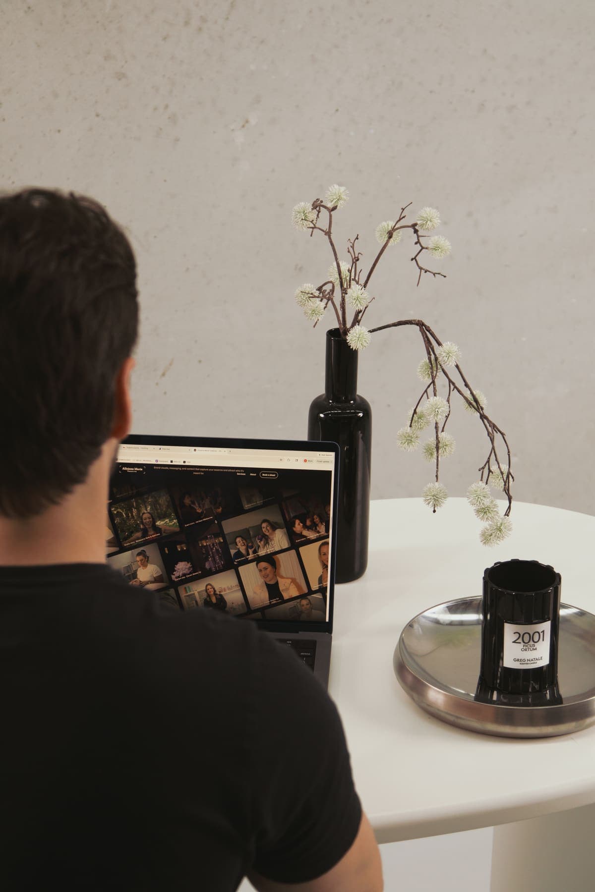 Over-the-shoulder view of Luke Marinovic at his laptop, a grid of photos on screen, vase of blossom on the desk.