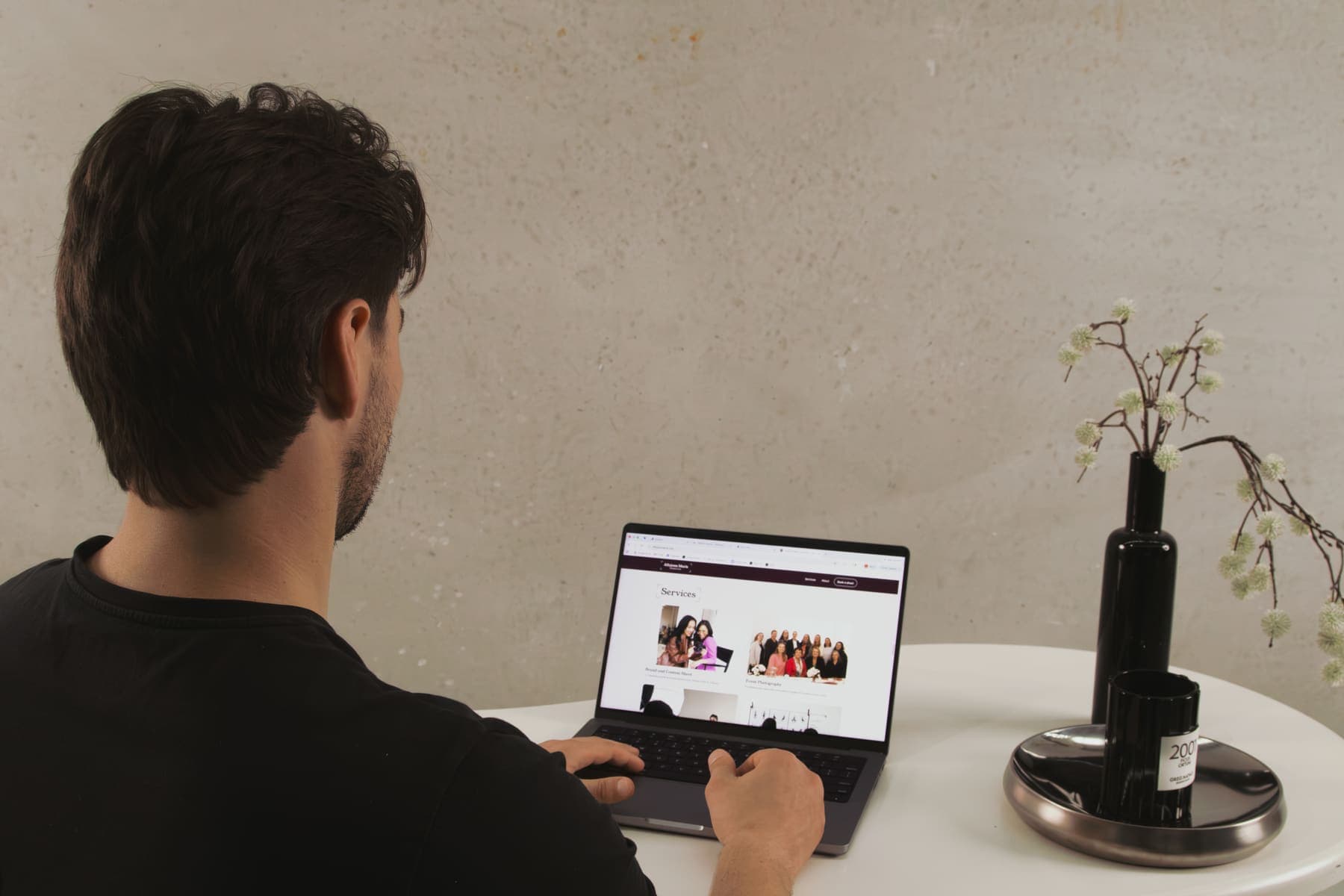 Luke Marinovic seen from behind, typing on a laptop at a round white table, a website open on screen.