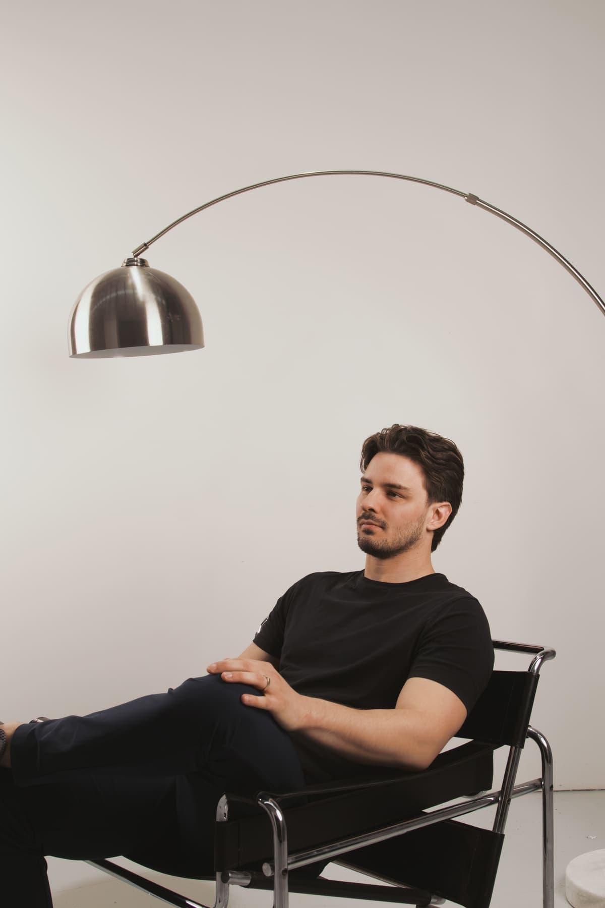 Luke Marinovic relaxed in a black sling chair, hands folded, under a chrome arc lamp.