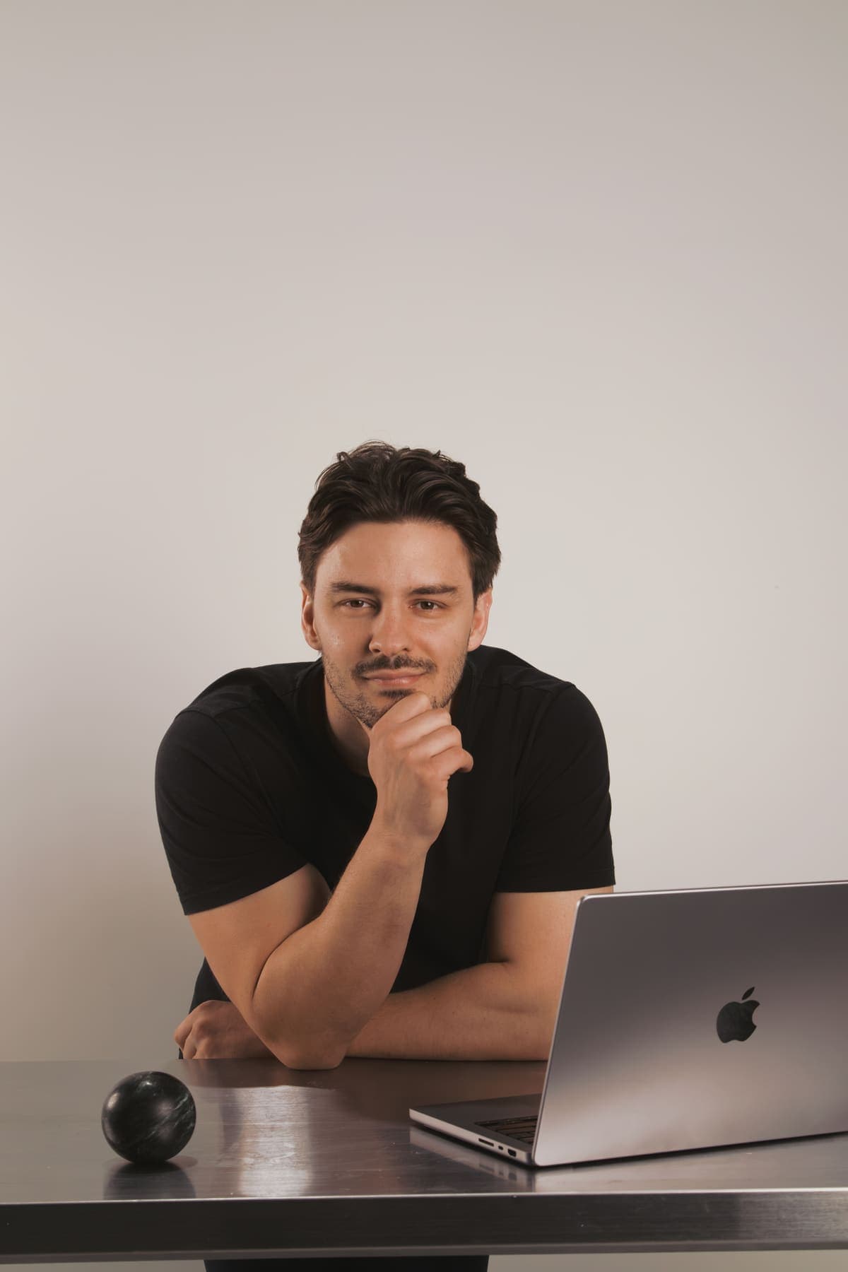 Luke Marinovic smiling with his hand on his chin at a steel desk, laptop open.