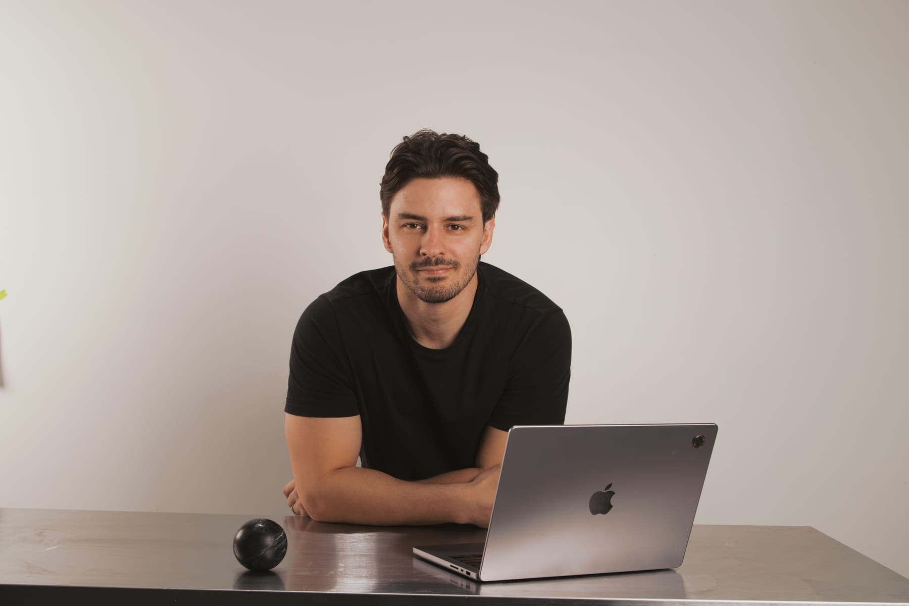 Luke Marinovic leaning on a steel desk beside an open laptop, looking at the camera.