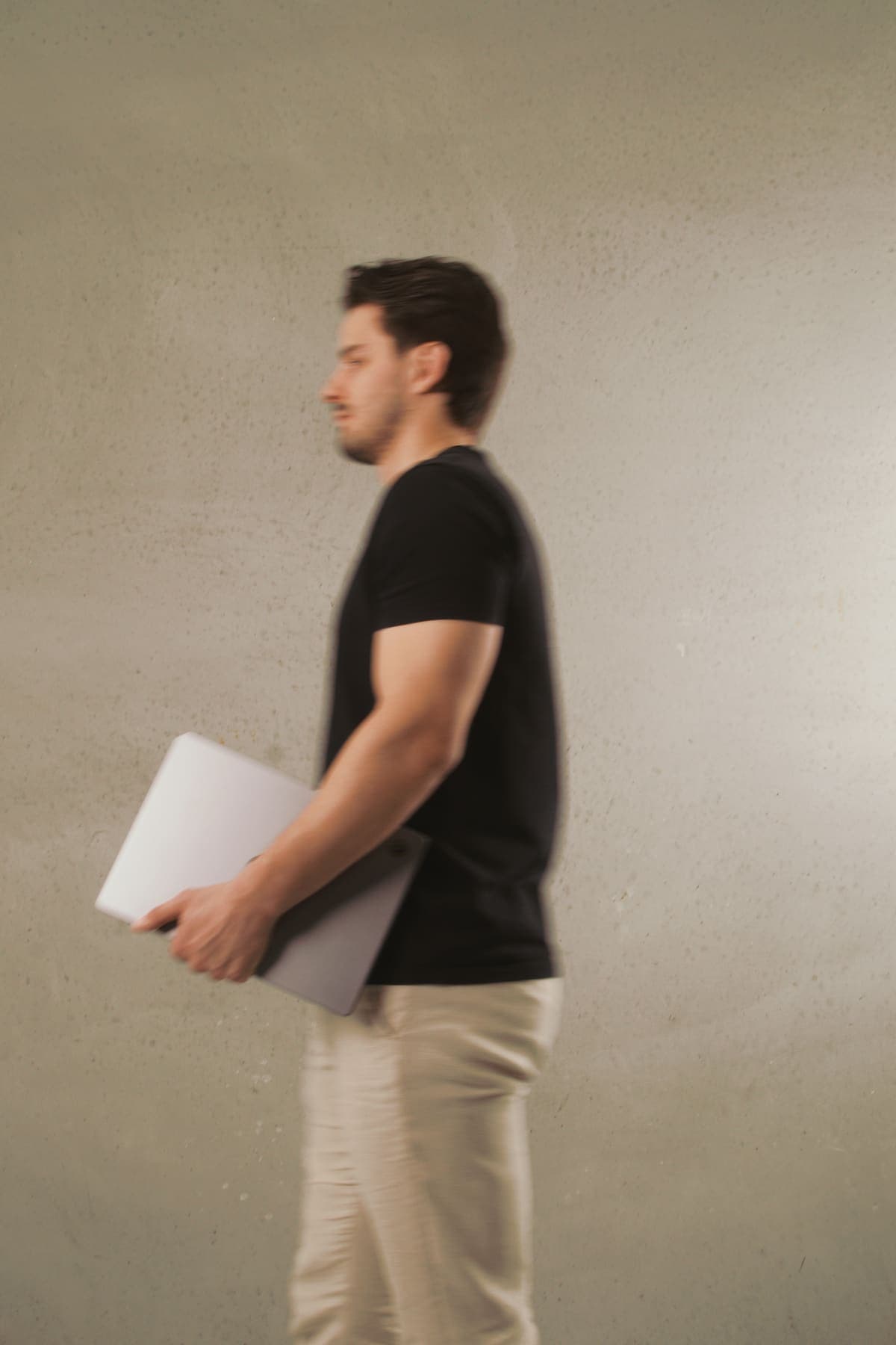 Luke Marinovic walking in profile with a laptop under his arm, past a concrete wall.