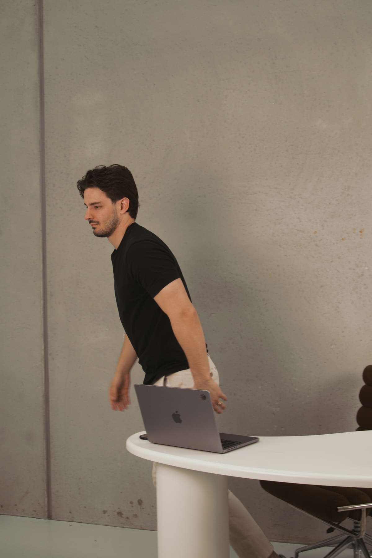 Luke Marinovic walking past his desk towards an open laptop, in a black t-shirt against a concrete wall.
